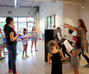 Girls gathered around a piano with sheet music singing. Katie Kolczun